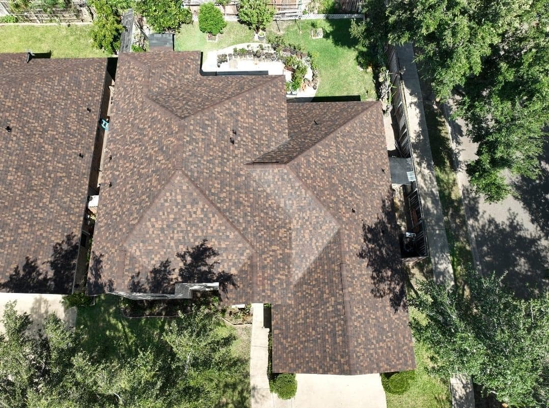 Aerial view of a residential roof with brown shingles and surrounding greenery.